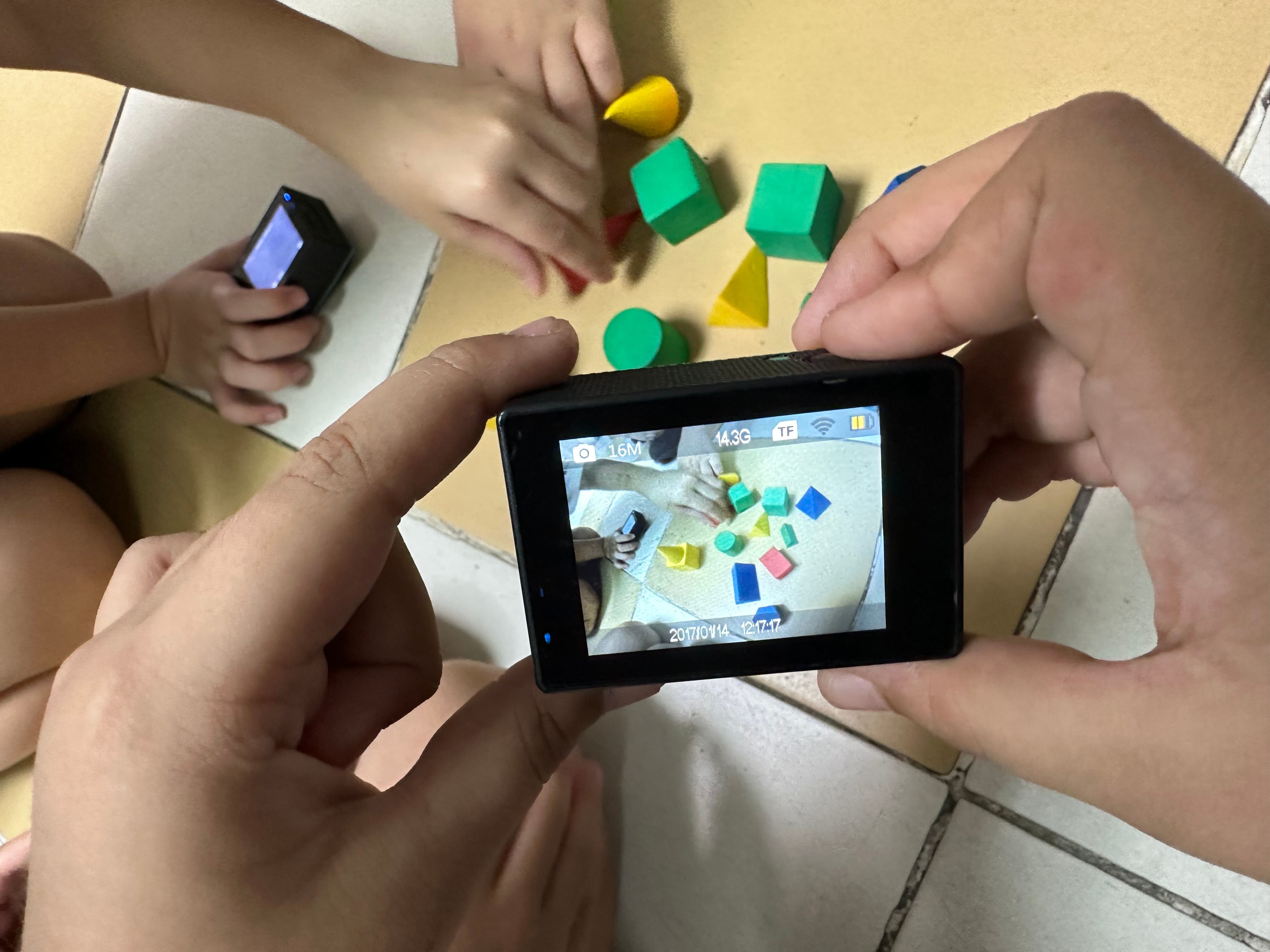 A hand holds a small camera filming a child playing with colorful wooden blocks on a tiled floor.